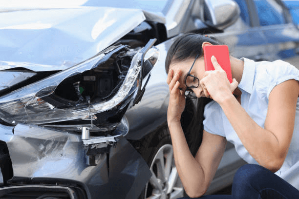 A woman kneels beside a damaged car while she makes a phone call and puts one hand against her forehead.