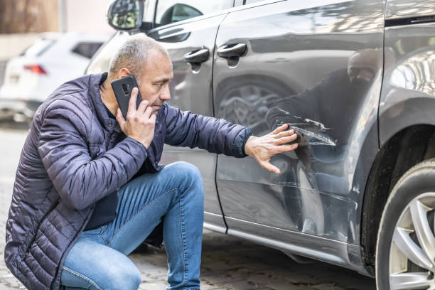 A man kneels beside a gray car to inspect the damaged door while talking on the phone.